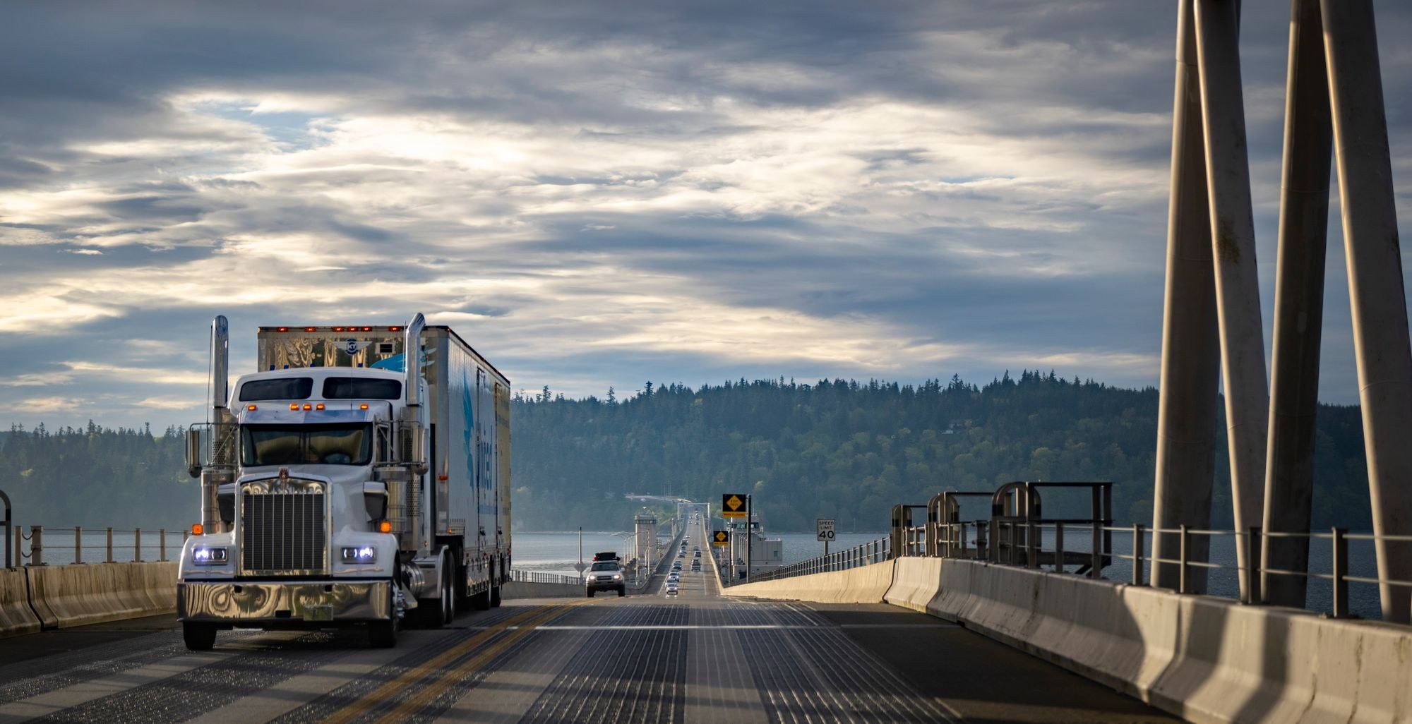 Silver American semi truck traveling across a bridge under a cloudy sky